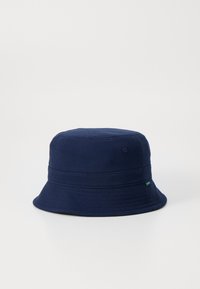 Chapeau seau bleu marine avec une texture en coton doux, couronne arrondie et large bord incliné vers le bas. Présente une petite étiquette verte sur le côté.