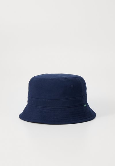 Chapeau seau bleu marine avec une texture en coton doux, couronne arrondie et large bord incliné vers le bas. Présente une petite étiquette verte sur le côté.