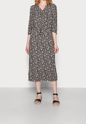 Kvinde iført en sort midi-kjole med små hvide og pink blomsterprint og sorte ankelstroppesandaler, stående foran en ensfarvet baggrund.