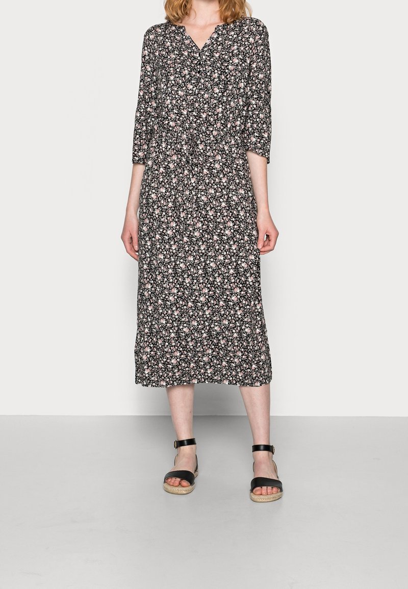 Femme portant une robe midi noire avec un petit imprimé floral blanc et rose, et des sandales noires à bride de cheville, debout devant un fond uni.