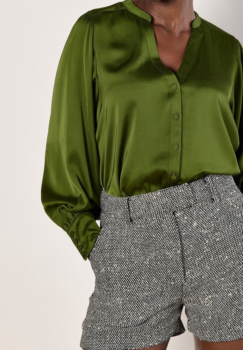 Femme portant un chemisier boutonné vert olive brillant à manches longues et un short gris texturé taille haute avec poches.