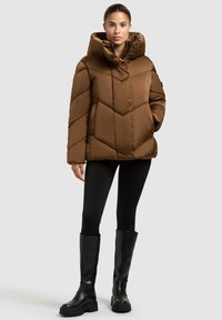 Giubbotto puffo marrone con colletto alto, caratterizzato da un design trapuntato e un ampio cappuccio. Indossato con leggins neri e stivali neri fino al ginocchio.