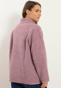 Chaqueta de forro polar de color púrpura claro con un alto cuello, superficie texturizada y hombros caídos. Presenta un corte relajado y un dobladillo curvado.