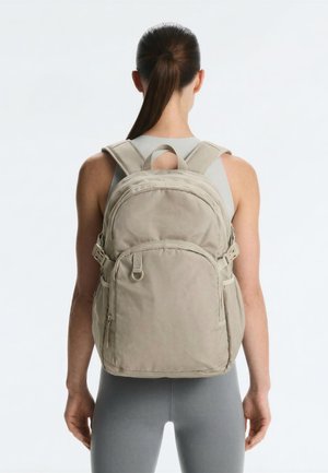 Person mit dunklen Haaren im Pferdeschwanz, die einen beigen Rucksack mit mehreren Reißverschlussfächern trägt, steht vor einem einfarbigen weißen Hintergrund.