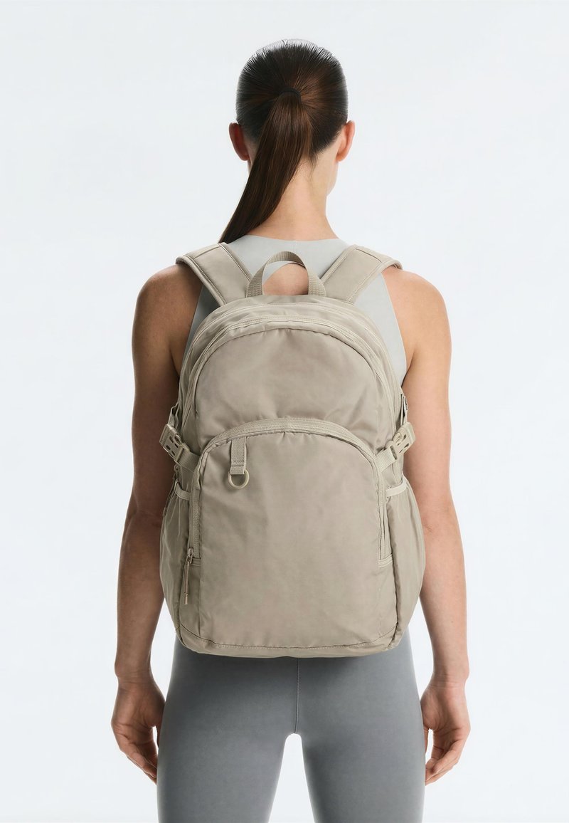 Personne aux cheveux foncés en queue de cheval, portant un sac à dos beige avec plusieurs compartiments zippés, debout devant un fond blanc uni.