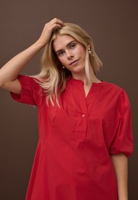 Femeie blondă purtând cercei cu perle și o rochie roșie lejeră, atingându-și părul pe un fundal maro simplu.