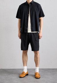 Zwarte t-shirt met korte mouwen, witte gestructureerde top eronder, zwarte cargoshorts en geweven bruine instappers met witte sokken.