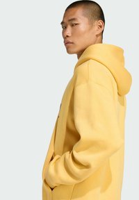 Gelber Kapuzenpullover aus weichem Stoff, mit großer Fronttasche und Kordelzugkapuze, von der Seite an einer Person dargestellt.