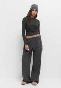 Graues Langarmoberteil mit gerafftem Stoff und passender locker geschnittener Hose. Das Model trägt eine graue Mütze und die Schuhe haben eine hellbraune Farbe.
