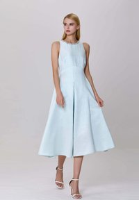 Une robe midi sans manches bleu clair en tissu lisse, avec une jupe plissée et un col en perles. Des talons blancs à lanières complètent le look.