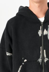 Black zip-up hoodie featuring a soft texture, oversized fit, and white paint splatter accents on the front and pocket.