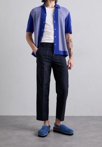 Koszula z krótkim rękawem w kolorze niebieskim i jasnoniebieskim, biały top na ramiączkach, granatowe spodnie z przeszyciami na kieszeniach, niebieskie chodaki z brązowym akcentem.