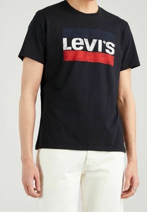 Mężczyzna w czarnej koszulce Levi's z białym i czerwonym logo oraz kremowych spodniach, stojący na jednolitym tle.