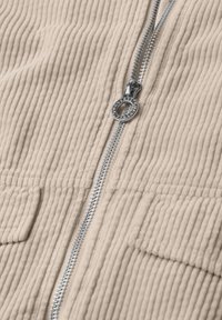 Close-up of beige corduroy fabric with vertical ridges and a silver metal zipper running down the center with rectangular pockets on each side.