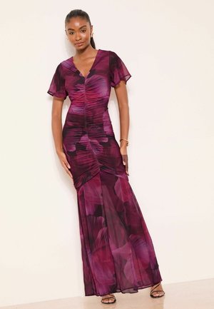 Kvinde iført en gulvlængde, tætsiddende lilla og sort blomsterkjole med korte flæseærmer, stående op ad en ensfarvet væg.