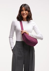 Borsa in tessuto viola di forma arrotondata con tracolla lunga, appoggiata su una blusa bianca e pantaloni grigi plissettati. Texture liscia, design minimale.