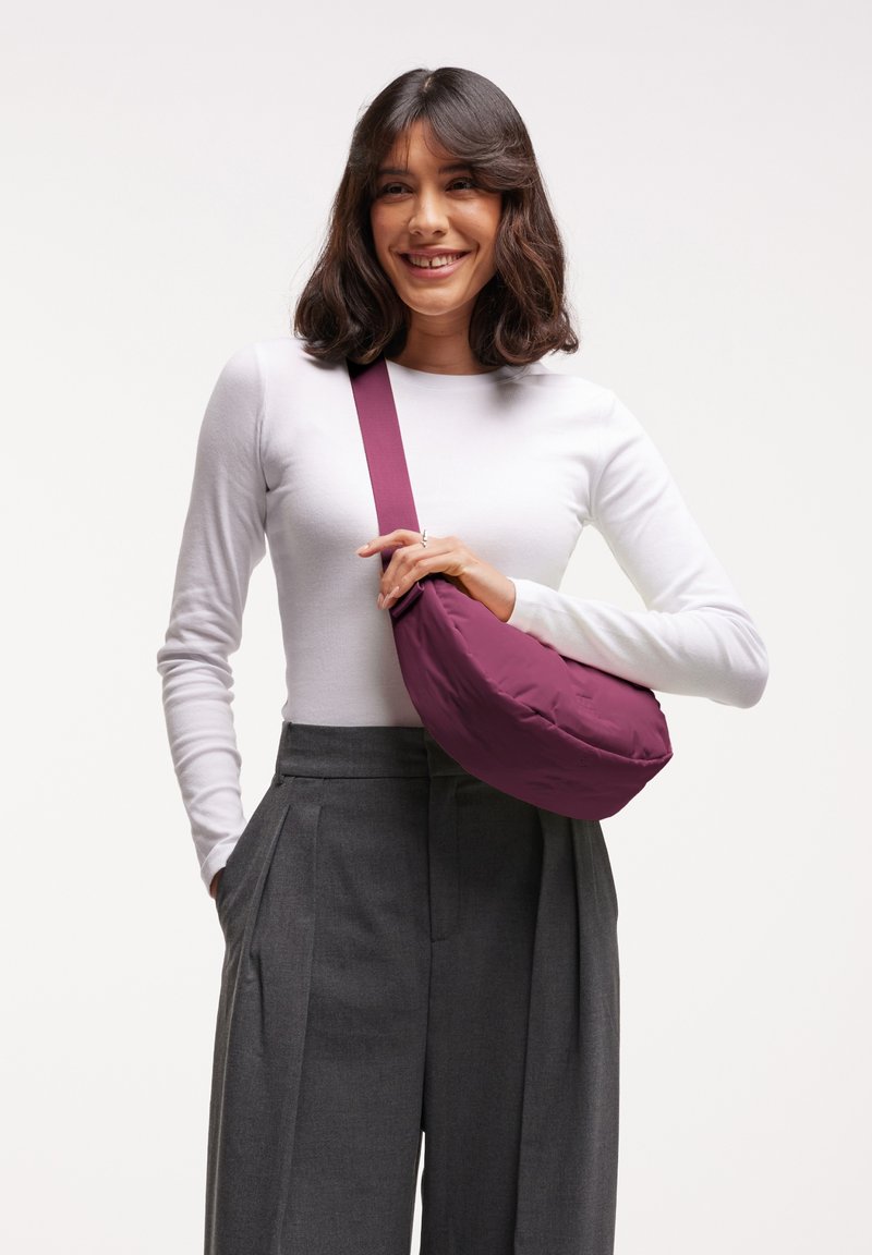 Borsa in tessuto viola di forma arrotondata con tracolla lunga, appoggiata su una blusa bianca e pantaloni grigi plissettati. Texture liscia, design minimale.