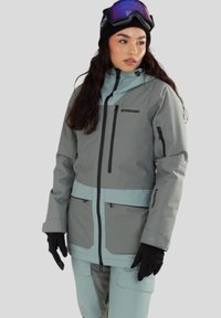 Grijze en mintgroene skijas met ritszakken en verstelbare manchetten, gedragen met een zwarte beanie en skibril. De stof heeft een gestructureerde afwerking.