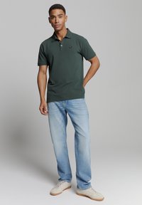 Donkergroen polo-shirt met korte mouwen, kraagontwerp en klein embleem; gecombineerd met lichtblauwe spijkerbroek en beige sneakers.