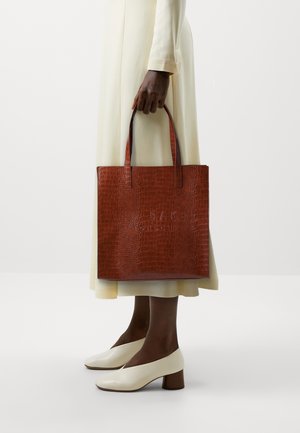 Brun texturerad handväska med krokodilmönster och två handtag upptill, hålls av en person. Mjuk klänning i krämfärg och vita blockklackade skor syns.