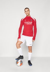 Piros, hosszú ujjú mez bordás textúrával és fehér díszítéssel, fehér nadrággal, amelyen fekete embléma található. Fekete cipők és focilabda.