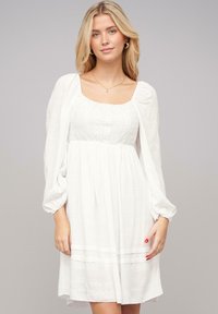 Vestido de algodón blanco con escote cuadrado, cuerpo texturizado, mangas abullonadas y una falda fluida. Presenta rayas horizontales en el dobladillo.