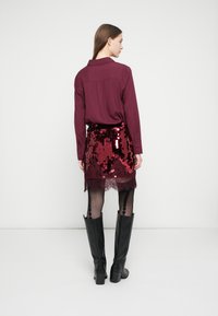 Kobieta zwrócona tyłem, ubrana w bordową bluzkę z długim rękawem, spódnicę z cekinami z koronkowym dołem, rajstopy w wzory oraz czarne kozaki za kolano.