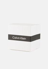 Valge ristkülikukujuline kast sileda tekstuuriga. Must riba, millel on valgega trükitud "Calvin Klein". Minimalistlik disain, ilma lisamustriteta.