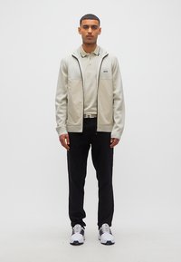 Άνδρας που στέκεται φορώντας ανοιχτό μπεζ ζακέτα με φερμουάρ, μπεζ πόλο πουκάμισο, μαύρο παντελόνι και λευκά αθλητικά παπούτσια μπροστά σε απλό λευκό φόντο.