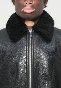 Veste en cuir noir avec une texture froissée, dotée d'un col en shearling noir, d'une fermeture éclair à l'avant et d'une coupe ajustée.