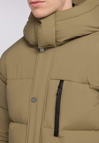 Giacca imbottita beige con colletto alto, ampie sezioni trapuntate, chiusura centrale con bottoni e una tasca nera con cerniera sul lato sinistro.
