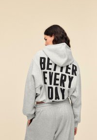 Grå hættetrøje med snøre i hætten og oversized pasform. Sort fed tekst "BETTER EVERY DAY" på ryggen. Blødt stof og afslappet design.