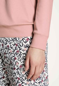 Light pink sweatshirt with ribbed cuffs, paired with white pants featuring dark leaves and pink floral patterns. Textured fabric details visible.