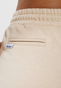 Los pantalones de chándal beige presentan una cinturilla acanalada y un bolsillo trasero ajustado. La tela es suave con una textura lisa, y se puede ver una pequeña etiqueta.