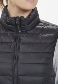 Chaleco aislante negro con cuello alto y cierre de cremallera, que presenta un patrón acolchado y un logotipo en el pecho. Textura de tela suave.