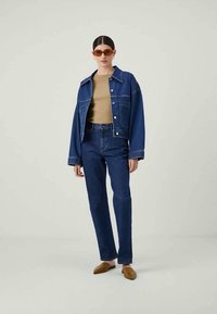 Mulher com cabelo apanhado, a usar óculos de sol castanhos, uma blusa bege, um casaco de denim azul oversized e jeans, e sapatos planos castanhos com bico fino.