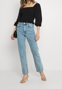 Haut noir à encolure carrée et smocké avec manches longues bouffantes ; jean bleu clair taille haute à jambes droites ; talons dorés avec lanières ; sac à main noir.