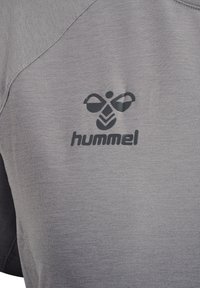 Szary materiał z czarnym logo Hummel przedstawiającym stylizowaną pszczołę nad nazwą marki na elemencie odzieży.