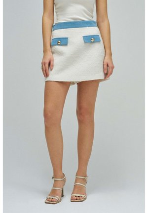 Frau trägt einen cremefarbenen Minirock mit Textur, blauem Bund und Taschenklappen, kombiniert mit weißen, mit Riemchen versehenen Sandaletten mit Absatz.