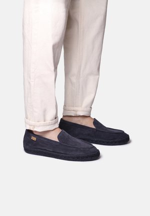 Zapatillas de ante azul marino sin cordones, con un superior texturizado y suela de tela trenzada, combinadas con pantalones crema remangados.