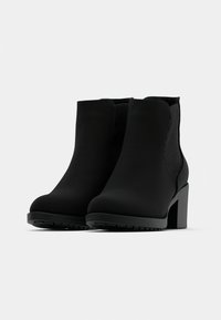 Un par de botas negras hasta el tobillo con tacones cuadrados y paneles laterales elásticos sobre un fondo blanco.