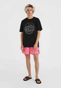 Camiseta negra con texto gráfico blanco y olas, combinada con bañador rosa con rayas horizontales y sandalias negras tipo slide.