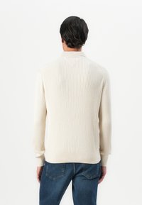 Homme aux cheveux foncés portant un pull crème à côtes et un jean bleu, se tenant de dos devant un fond blanc uni.