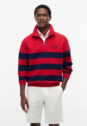 Man draagt een rood- en marineblauw gestreepte kwart-ritspullover met wit overhemd met kraag en witte korte broek, staand met één hand in de zak.