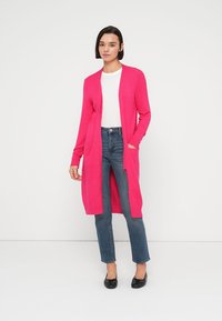 Ροζ μακρύ ζακέτα με ανοιχτό μπροστινό μέρος, ριμπ μανσέτες και δύο μπροστινές τσέπες, συνδυασμένη με στενά μπλε τζιν και μαύρα μπαλαρίνα.