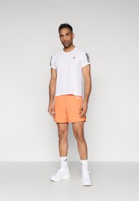 Λευκό αθλητικό t-shirt με μαύρες ρίγες στα μανίκια, συνδυασμένο με φωτεινά πορτοκαλί σορτς. Το μοντέλο φοράει λευκά αθλητικά παπούτσια και εμφανείς λευκές κάλτσες.