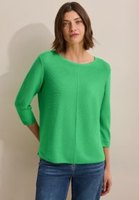 Haut en tricot vert avec un col rond, des manches trois-quarts et une coupe décontractée. Texture remarquable avec des détails côtelés et des fentes latérales.