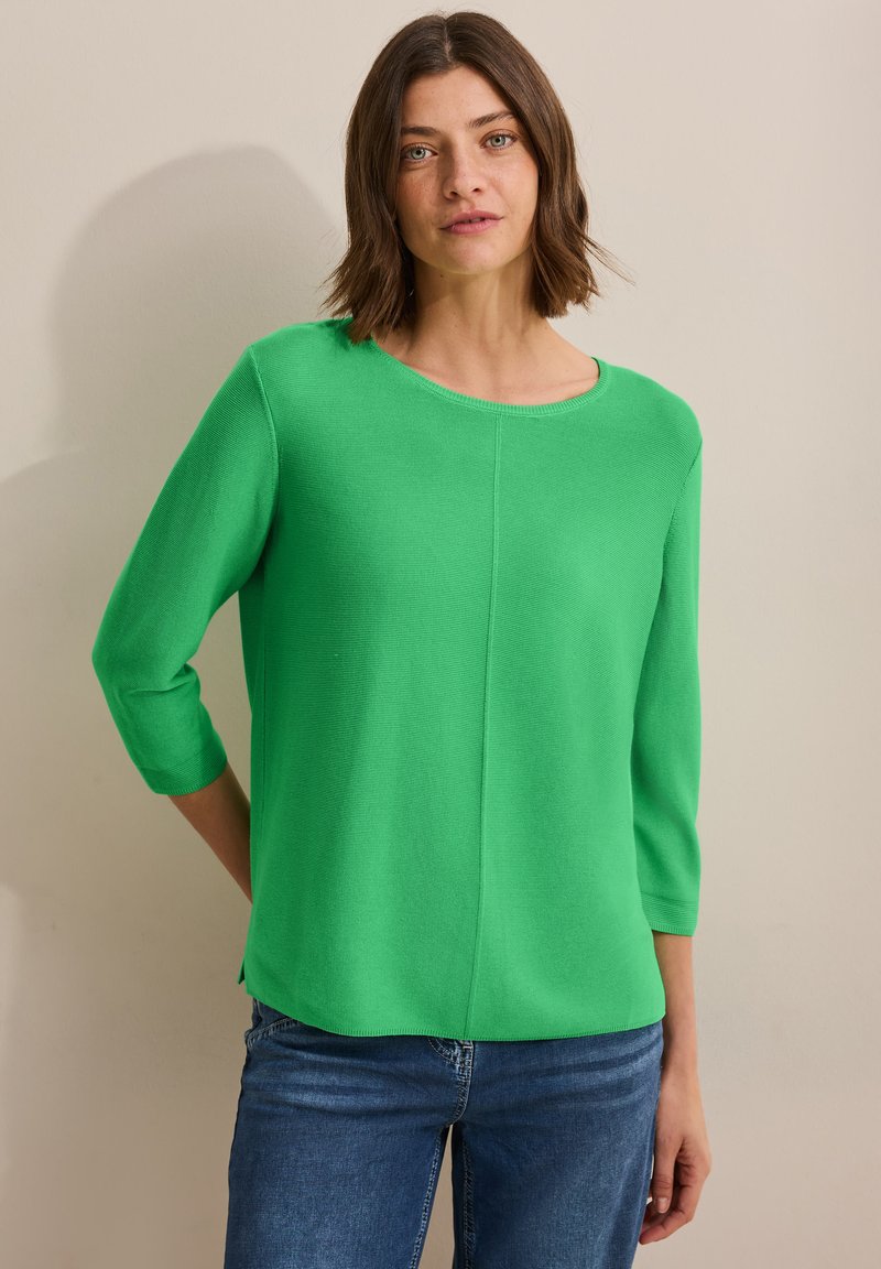 Grüner Strickoberteil mit rundem Ausschnitt, drei Viertelärmeln und lockerem Schnitt. Auffällige Textur mit gerippten Details und seitlichen Schlitzen.