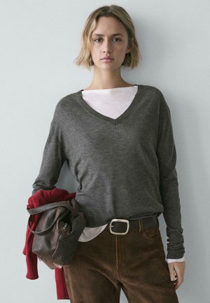 Gray v-neck sweater layered over a white shirt, paired with brown corduroy pants. Holding a brown bag and a red sweater.