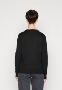 Svart stickad tröja med långa ärmar och rundad halsringning, med ribbat nederkant och ärmslut. Bärs över grå jeans. Visar bakifrån.